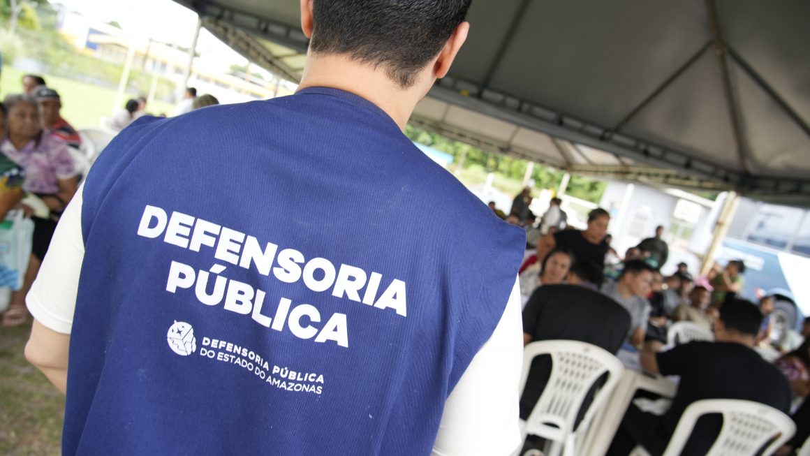 Homem de costas, utilizando um colete com os dizeres DEFENSORIA PÚBLICA e a logo da Defensoria Pública do Estado do Amazonas abaixo das palavras, participando de um mutirão de atendimentos.