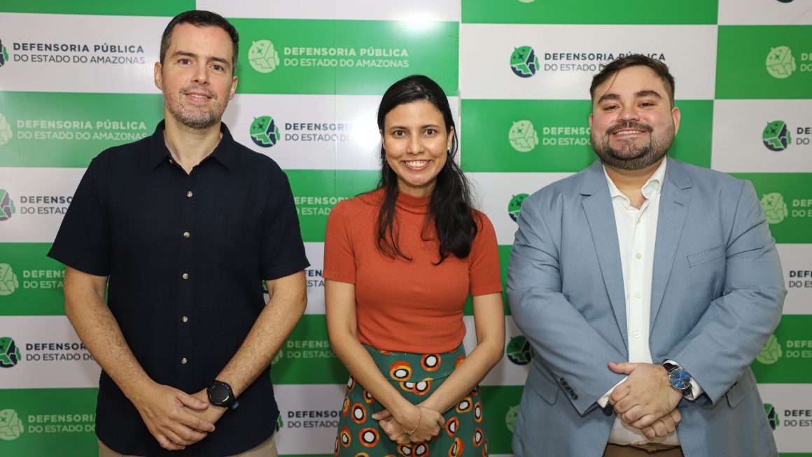 Na imagem, vemos os defensores Inácio Navarro, Karoline Santos e Lucas Matos em pé, posando para a foto, em frente ao backdrop da Defensoria, disposto no auditório da sede administrativa