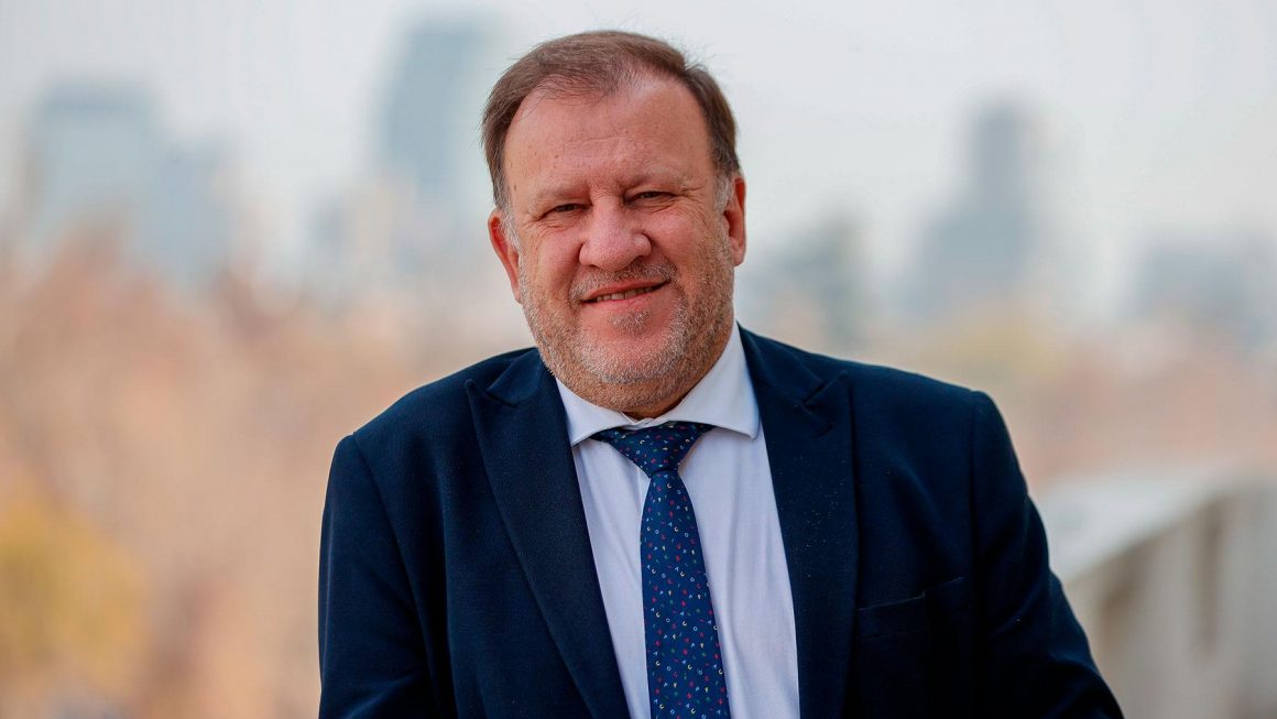 Professor Lorenzo Bujosa sorrindo usando terno e gravata azul escuro, quase preto, com uma paisagem de fundo desfocada