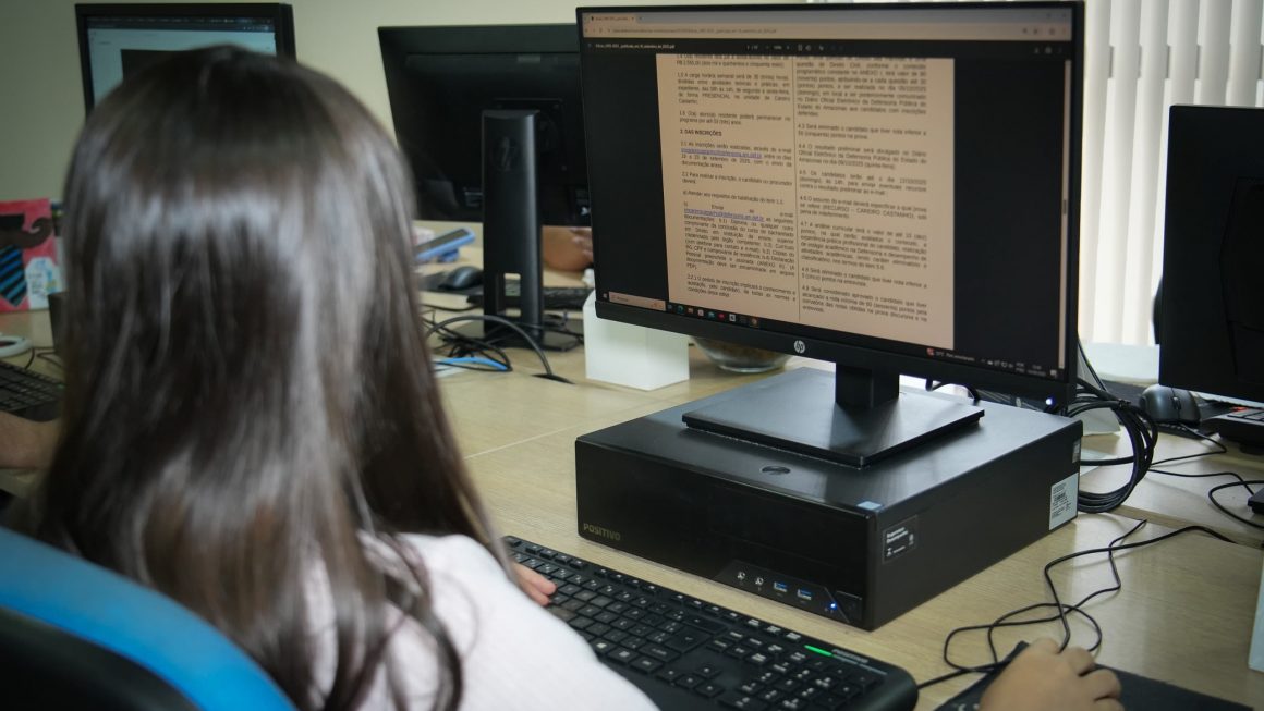 Estagiária de costas para a câmera visualiza o edital de inscrição na tela de um computador.
