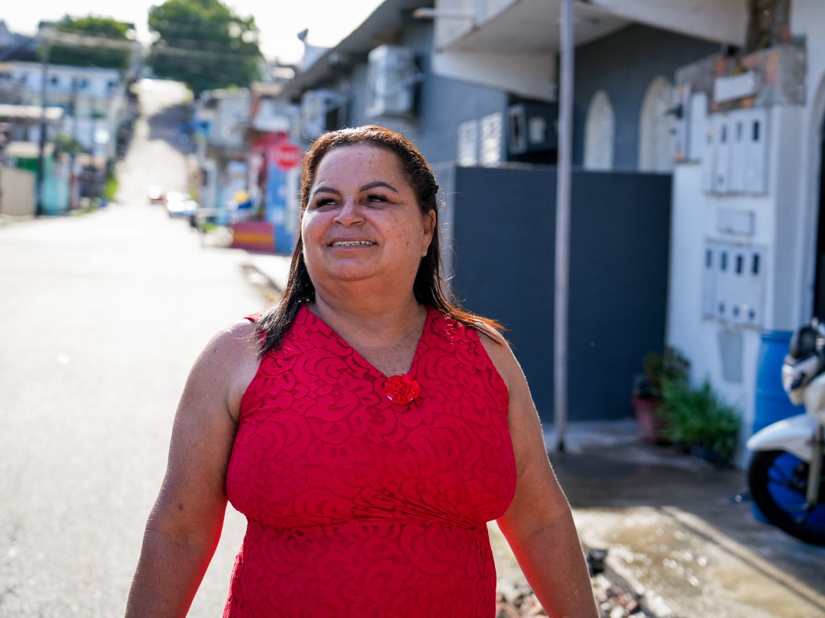 Na imagem, vemos Eliana Cristina, personagem principal da matéria; ela está vestida de vermelho, com um leve sorriso no rosto, olhando para o infinito, em uma tarde ensolarada, em frente a sua casa, em Manaus