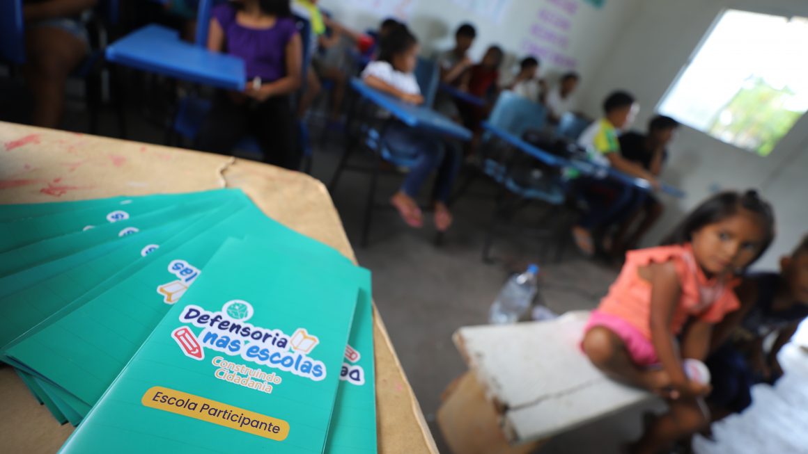 Na imagem vemos uma sala de aula, em primeiro plano um folder do projeto Defensoria nas Escolas, com uma menina ao fundo