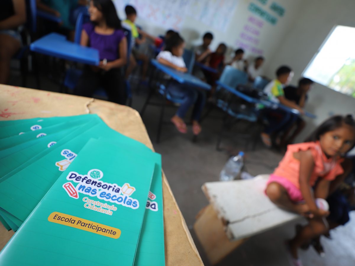 Na imagem vemos uma sala de aula, em primeiro plano um folder do projeto Defensoria nas Escolas, com uma menina ao fundo