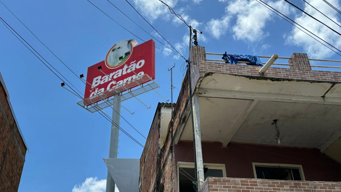 A imagem mostra a placa do supermercado Baratão da Carne ao lado de uma casa afetada por uma obra da empresa