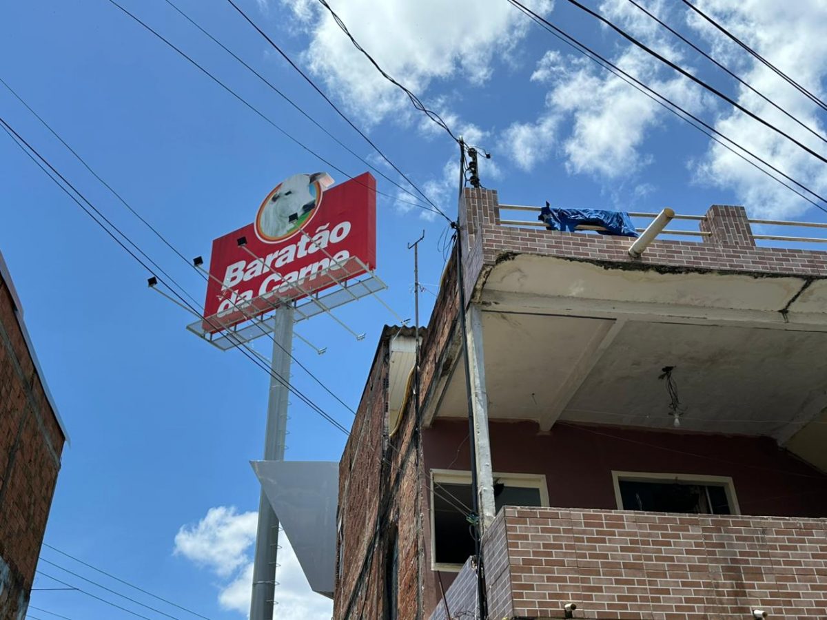 A imagem mostra a placa do supermercado Baratão da Carne ao lado de uma casa afetada por uma obra da empresa