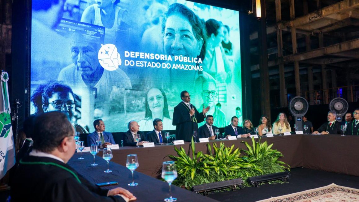 Imagem mostra a mesa de autoridades durante a posse, com o defensor geral em pé ao centro, com microfone na mão