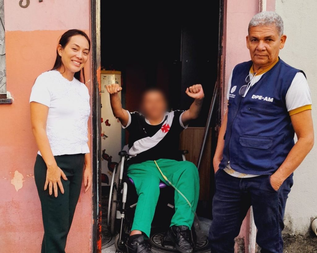 Na imagem vemos o asssistido cadeirante em sua cadeira de rodas ao lado da defensora do caso e um servidor da DPE-AM, todos posando para a foto. O assistido está com o rosto borrado e com os braços levantados