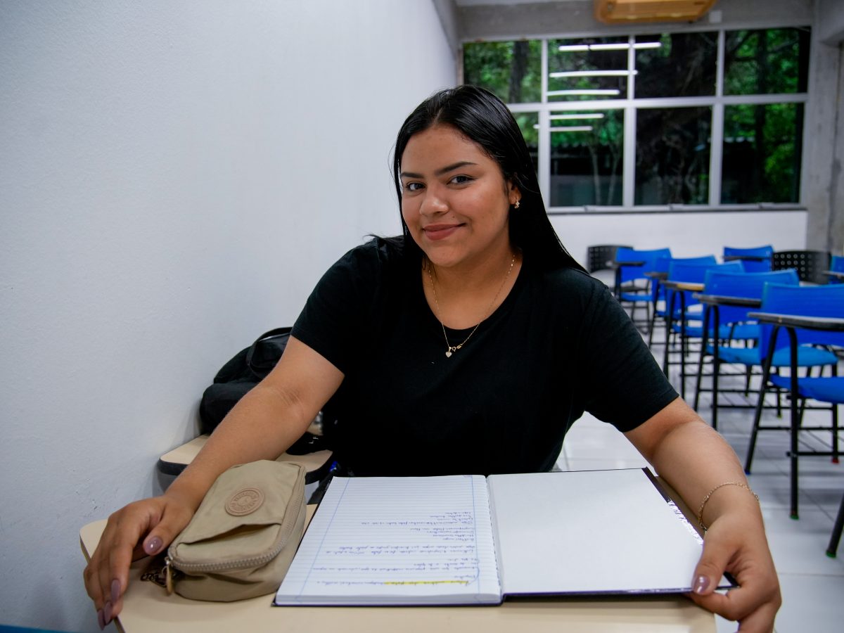A foto mostra Emanuelle Õkapa, uma jovem mulher de longos cabelos pretos, vestindo uma camiseta preta, sentada em uma escrivaninha de sala de aula da Ufam e sorrindo para a câmera. Ela está posicionada na frente de um caderno aberto e segura um estojo de lápis de tecido bege. O fundo mostra uma sala de aula com várias fileiras de cadeiras azuis vazias e janelas com vista para árvores.