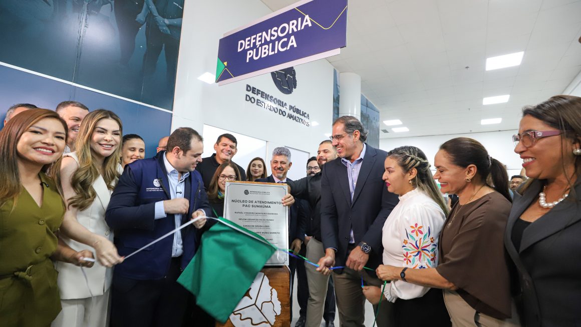 Uma fotografia da cerimônia de inauguração da nova unidade da Defensoria Pública do Estado do Amazonas (DPE-AM) no PAC Sumaúma. O Governador Interino do Amazonas, Roberto Cidade (segundo à esquerda, vestindo paletó azul), e o Defensor Público-Geral (DPG), Rafael Barbosa (terceiro à esquerda, vestindo paletó preto e óculos), estão no centro da imagem, prestes a cortar a fita inaugural verde e branca. Eles são ladeados por uma equipe de apoio e convidados. Em primeiro plano, uma placa de inauguração coberta com tecido verde e montada em uma base de madeira com o logotipo da DPE e a inscrição "DPE" está visível. Acima do grupo, letreiros pendurados e de parede exibem o texto "DEFENSORIA PÚBLICA" e "DEFENSORIA PÚBLICA DO ESTADO DO AMAZONAS". O ambiente é um espaço interno de serviço público moderno e bem iluminado.
