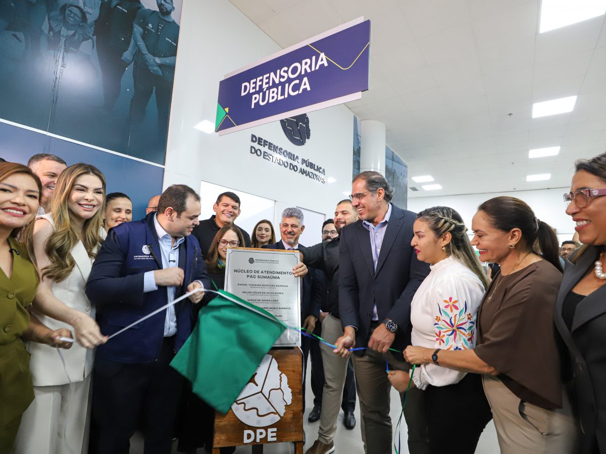 Uma fotografia da cerimônia de inauguração da nova unidade da Defensoria Pública do Estado do Amazonas (DPE-AM) no PAC Sumaúma. O Governador Interino do Amazonas, Roberto Cidade (segundo à esquerda, vestindo paletó azul), e o Defensor Público-Geral (DPG), Rafael Barbosa (terceiro à esquerda, vestindo paletó preto e óculos), estão no centro da imagem, prestes a cortar a fita inaugural verde e branca. Eles são ladeados por uma equipe de apoio e convidados. Em primeiro plano, uma placa de inauguração coberta com tecido verde e montada em uma base de madeira com o logotipo da DPE e a inscrição "DPE" está visível. Acima do grupo, letreiros pendurados e de parede exibem o texto "DEFENSORIA PÚBLICA" e "DEFENSORIA PÚBLICA DO ESTADO DO AMAZONAS". O ambiente é um espaço interno de serviço público moderno e bem iluminado.