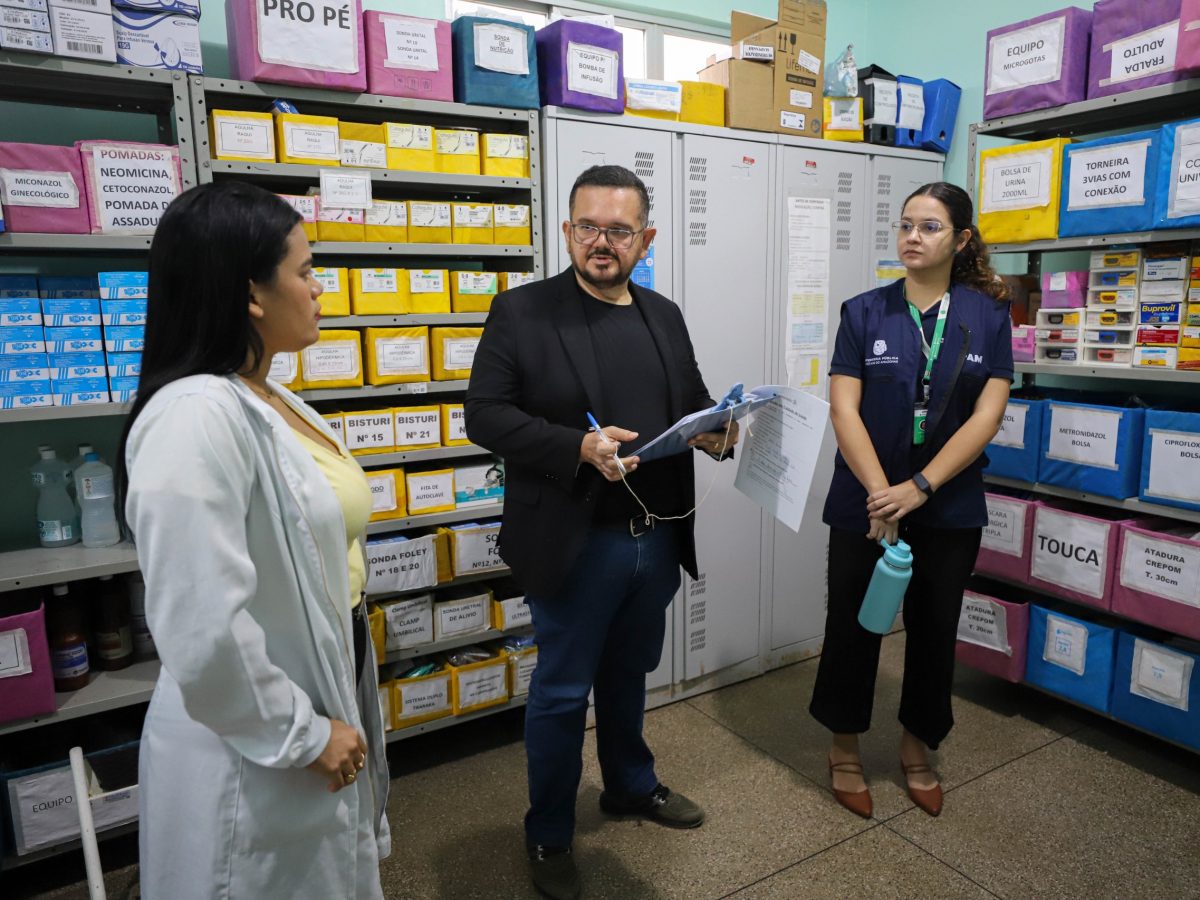 Na imagem, vemos o defensor Arlindo Gonçalves, ao centro, com uma prancheta na mão. Ao seu lado esquerdo está uma assessora e, do lado direito, uma profissional da saúde de jaleco; os três estão dentro de um depósito de medicamentos de um hospital