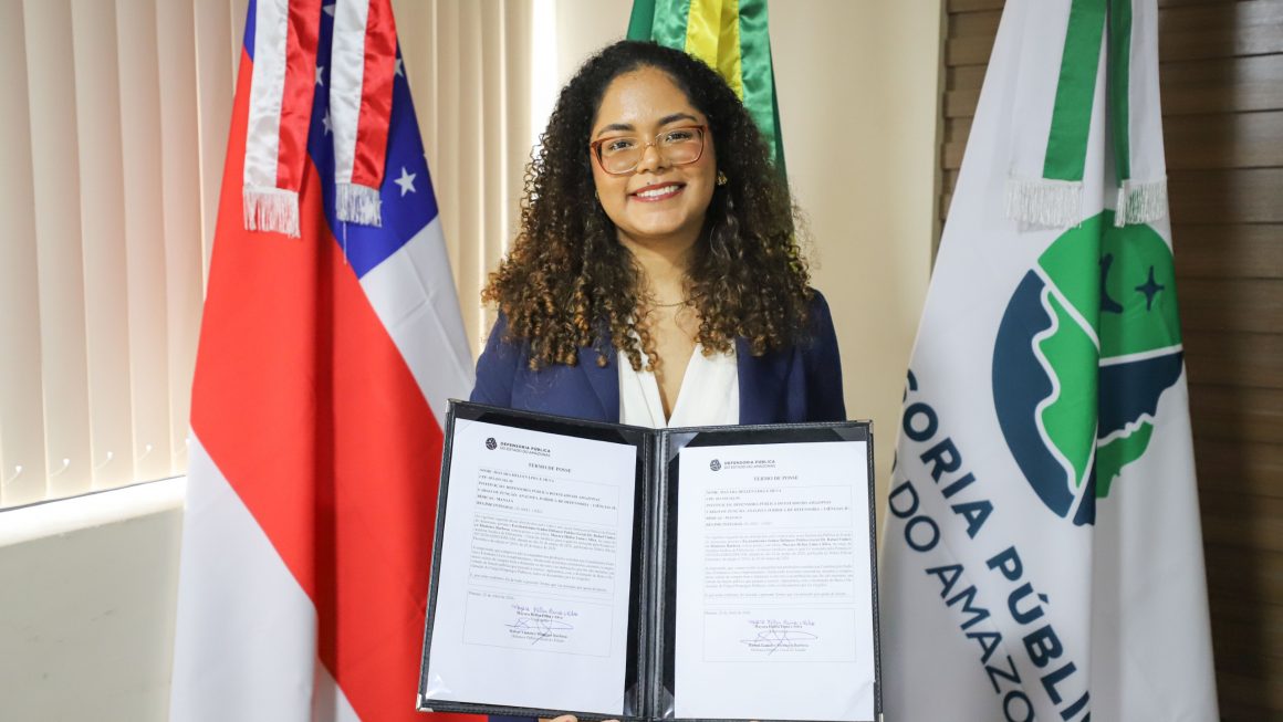 Uma mulher jovem, de cabelos cacheados, sorri para a foto enquanto segura um porta-diploma aberto com o documento de posse. Ela usa óculos de armação clara e um blazer azul marinho sobre uma blusa branca. Ao fundo, estão posicionadas três bandeiras: a do estado do Amazonas à esquerda, a do Brasil ao centro e a bandeira branca da Defensoria Pública do Estado do Amazonas (DPE-AM) à direita. O ambiente é formal e bem iluminado.