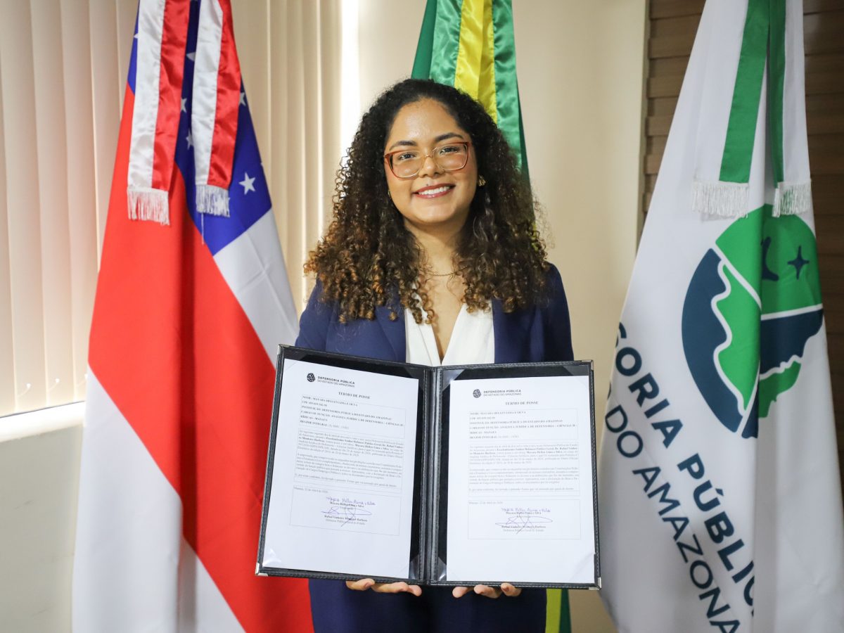 Uma mulher jovem, de cabelos cacheados, sorri para a foto enquanto segura um porta-diploma aberto com o documento de posse. Ela usa óculos de armação clara e um blazer azul marinho sobre uma blusa branca. Ao fundo, estão posicionadas três bandeiras: a do estado do Amazonas à esquerda, a do Brasil ao centro e a bandeira branca da Defensoria Pública do Estado do Amazonas (DPE-AM) à direita. O ambiente é formal e bem iluminado.